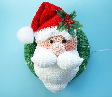 Gehäkelter Türkranz mit Weihnachtsmann-Gesicht, roter Mütze, weißem Bart, Stechpalmenzweig und kleiner Lichterkette vor hellblauem Hintergrund.