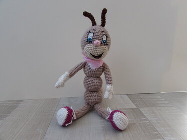 Gehäkelte Amigurumi-Raupe mit rosa Halstuch, blauen Augen und magenta-weißen Füßen, sitzend auf Holzfußboden.