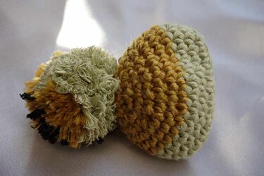 Gehäkelter eiförmiger Eierwärmer in Grün und Gelbbraun mit flauschigem Pompon auf hellem Stoff