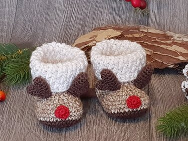Pair of crocheted reindeer baby booties with cream cuffs, brown antlers and red noses on a wooden floor
