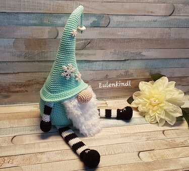 Sitzender, gehäkelter Wichtel mit mintgrüner Zipfelmütze, weißem Bart, beiger Nase und schwarz-weiß gestreiften Beinen, dekoriert mit Schneeflocken und kleinem Glöckchen.