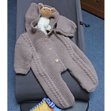 Taupefarbener gestrickter Baby-Overall mit Kapuze, Zopfmuster und drei Knöpfen; kleines Stofftier in der Kapuze