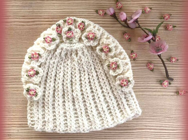 Cremefarbene gehäkelte Beanie mit rosa Blütenapplikationen auf hellem Holzhintergrund
