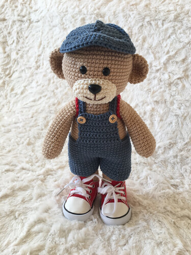 Gehäkelter beiger Teddybär in blauer Latzhose, Schirmmütze und roten Schnürschuhen auf weißem Fell
