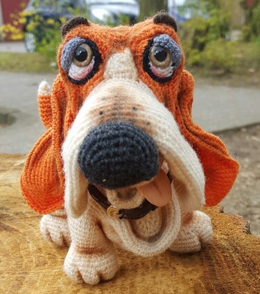 Gehäkelter Amigurumi Basset-Hund mit orange-braunen Schlappohren, großer schwarzer Nase, Halsband und herausgestreckter Zunge auf einem Holzstamm