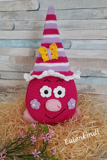 Gehäkelte Amigurumi-Dekofigur in Pink mit gestreiftem Hut und gelbem Schmetterling auf Stroh vor Holzverkleidung.