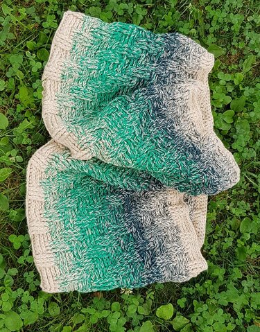 Gehäkelter Loop‑Schal mit Korbgeflechtmuster und Farbverlauf (Beige–Grün–Blau) auf Wiese