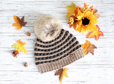 Braun-beige gehäkelte Bommelmütze mit Noppenmuster und Herzanhänger auf der Krempe, umgeben von Herbstblättern und Sonnenblume auf hellem Holz.