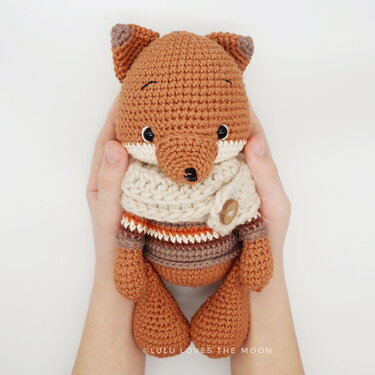 Hände halten einen braunen gehäkelten Amigurumi-Fuchs mit beigem Schal und Knopf