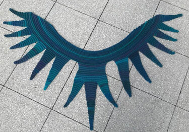 Halbmondförmiger gehäkelter Schal mit gezackten Spitzen in Blau- und Grüntönen auf grauen Bodenfliesen