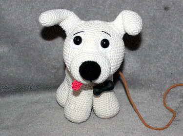 Gehäkeltes weißes Amigurumi-Hundchen mit schwarzen Augen, schwarzer Nase, roter Zunge und Knochenanhänger auf grauem Hintergrund