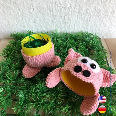 Nahansicht zweier rosa Häkel‑Schweinchen‑Hüllen mit gelben Innenkapseln auf grüner Deko