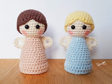Zwei gehäkelte Amigurumi-Engelchen in Rosa und Hellblau mit Flügeln und schwarzen Sicherheitsaugen auf Holz vor weißem Hintergrund.