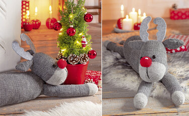 Gestricktes graues Rentier mit roter Nase liegt vor kleinem Weihnachtsbaum
