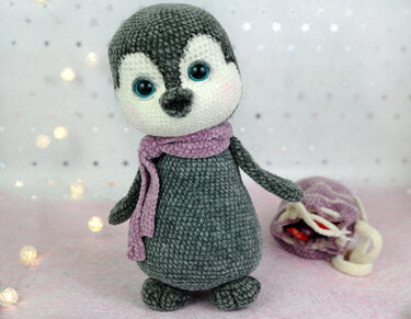 Gray crocheted penguin amigurumi wearing a lavender scarf on a pink surface