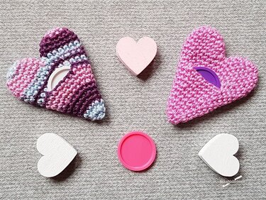 Zwei schnell gehäkelte Herzhüllen in Pink und Multicolor mit sichtbaren runden Kunststoffdeckeln und drei Holzherzen auf Textiluntergrund