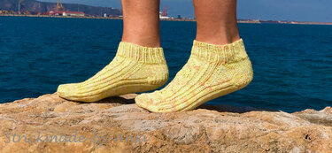Füße in gelben handgestrickten Knöchelsocken auf Felsen vor blauem Meer