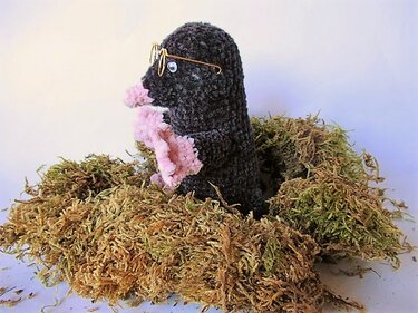 Gehäkeltes Maulwurf-Amigurumi mit Drahtbrille, das aus einem Moosnest herausblickt und rosa Schnauzen- und Pfotendetails zeigt