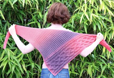 Person von hinten mit ausgestreckten Armen und einem gehäkelten Dreieckstuch in Pink‑Violett mit Rippenstruktur und Zopfmuster