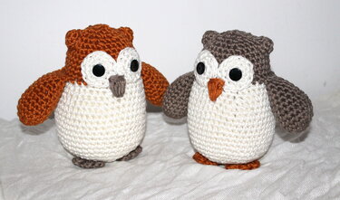 Zwei handgehäkelte Amigurumi-Eulen mit weißem Bauch, eine braun, eine orange, auf hellem Stoff