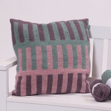 Knitted pillow with vertical block stripes in mauve, pink and green beside yarn balls