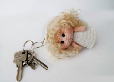 Crocheted amigurumi angel keychain with curly blonde hair and white dress attached to keys on a white background.