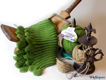 Halbfertiger Rippen-Kragenschal in Grün mit Zierkante, daneben Garnknäuel, Bindfaden und Strickzubehör