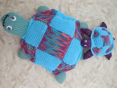 Zwei gestrickte Amigurumi-Schildkröten, groß und klein, mit hellblauen und mehrfarbigen Panzern und Wackelaugen auf hellem Hintergrund.