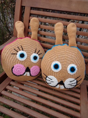 Zwei runde gehäkelte Hasen-Kissen mit langen Ohren, großen Augen, Schnurrhaaren und Zähnchen auf einer Holzbank