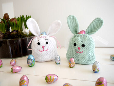 White and mint crocheted bunny bags with embroidered faces, filled with foil-wrapped eggs and surrounded by more eggs on a white surface.