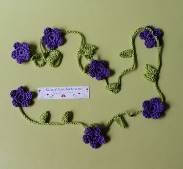 Gehäkelte Girlande mit lila Blumen und grünen Blättern auf hellgrünem Hintergrund.