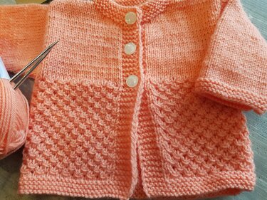 Apricotfarbenes gestricktes Babyjäckchen mit feinem Strukturmuster, drei hellen Knöpfen sowie Garn und Stricknadeln sichtbar