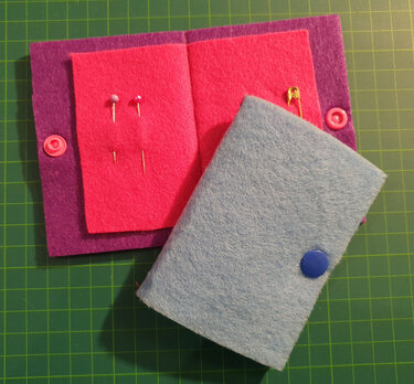 Offenes violettes Filzetui mit pinkem Nadelpad und Stecknadeln, daneben ein geschlossenes hellblaues Filzetui mit Druckknopf auf grüner Schneidematte