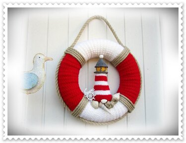 Gehäkelter rot-weißer Rettungsring mit kleinem Leuchtturm, Anker und Muscheln an weißer Bretterwand