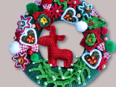 Handmade crochet Christmas wreath with a red reindeer, poinsettia flowers, holly leaves, bows, hearts, pom-poms, and small bells.