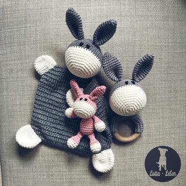 Grey crocheted security blanket with donkey head, wooden-ring rattle, and small pink plush on neutral fabric