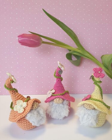Drei gehäkelte Wichtel in Pastellfarben mit Blumen und grünen Ranken vor rosa Hintergrund und einer Tulpe