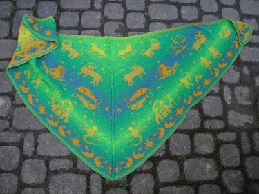 Gestricktes dreieckiges Tuch in Grün-Türkis mit gelb-orangenen Sternzeichen-Motiven liegend auf Kopfsteinpflaster