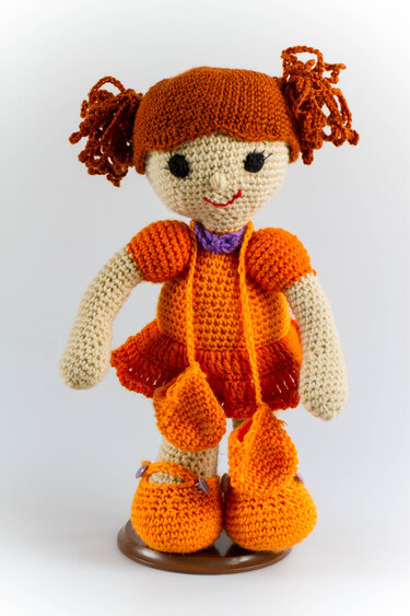 Handmade crocheted amigurumi doll with reddish-orange pigtails, orange dress, purple collar, and small matching pouches.