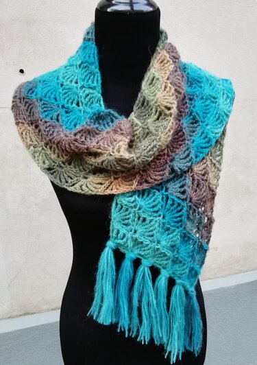 Gehäkelter Schal in melierten Türkis-, Grün- und Brauntönen mit Muschelmuster und Quasten auf einer schwarzen Schneiderbüste.