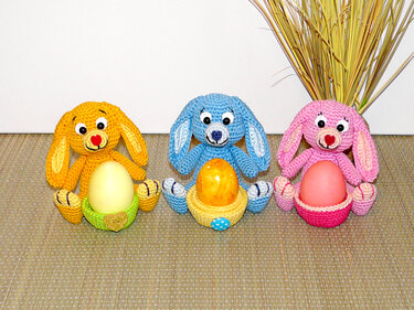 Three crocheted bunny egg holders in yellow, blue and pink, each holding a colored egg in a small basket