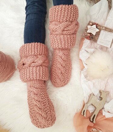 Altrosafarbene, gestrickte Socken und Beinstulpen mit Zopfmuster auf weißem, flauschigem Untergrund.