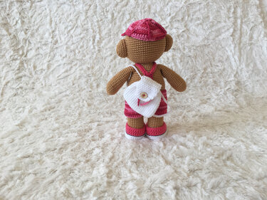 Gehäkelter brauner Teddybär von hinten mit rosa Mütze, weißem Rucksack mit Knopf und rosafarbenen Schuhen auf hellem, flauschigem Untergrund.