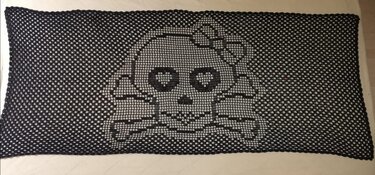 Rechteckiges schwarzes Filet-Häkeltuch mit Totenkopf- und gekreuzten Knochen-Motiv auf hellem Untergrund.