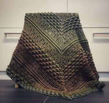 Ombre crochet blanket in green and brown tones with lace and textured stitch panels, draped over a stool.
