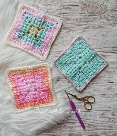 Close-up of three pastel textured crochet square swatches with a crochet hook and small embroidery scissors on wood and faux-fur surfaces