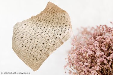 Hellbeige gestrickte Babydecke mit Rauten‑Lochmuster und kraus‑rechts gestricktem Rand auf hellem Hintergrund, daneben rosa Trockenblumen.