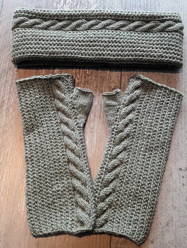 Grünes gestricktes Stirnband und fingerlose Handschuhe mit Zopfmuster auf Holzuntergrund.