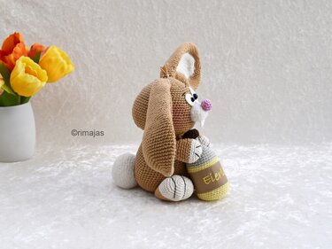 Gehäkelter Schlappohr-Hase in Braun mit rosa Nase und kleinem gehäkeltem Accessoire, Tulpen im Hintergrund