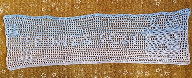 Rechteckiger gehäkelter Tischläufer mit Hellblau‑Weiß‑Farbverlauf und Lochmuster auf goldbraunem, sternverziertem Untergrund.